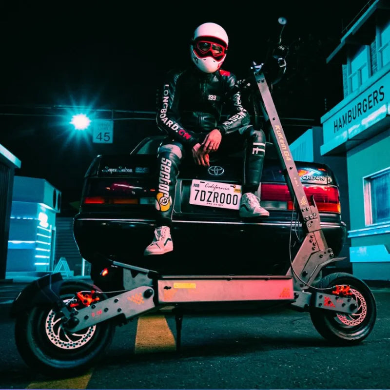 Guy in full motorcycle gear sitting on the boot of a car in the streets in the dark with a Kugoo KuKirin (Kugoo) G4 Electric Scooter in front of him.